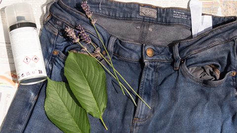 Eine alte Jeans wird mit einer Sprühdose besprüht. Auf der Hose liegen Blätter und Lavendel, darunter befindet sich eine Zeitung.